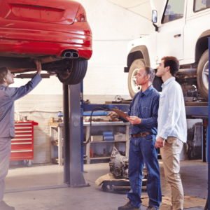 Three men stand in a garage with a car on a lift, discussing details related to an MOT tester course.