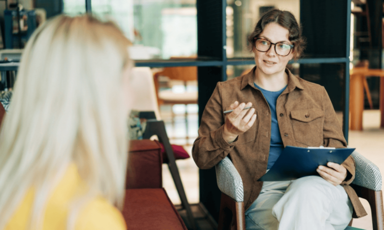 A woman speaks to another woman in a meeting room, discussing topics related to an online psychology conversion course.